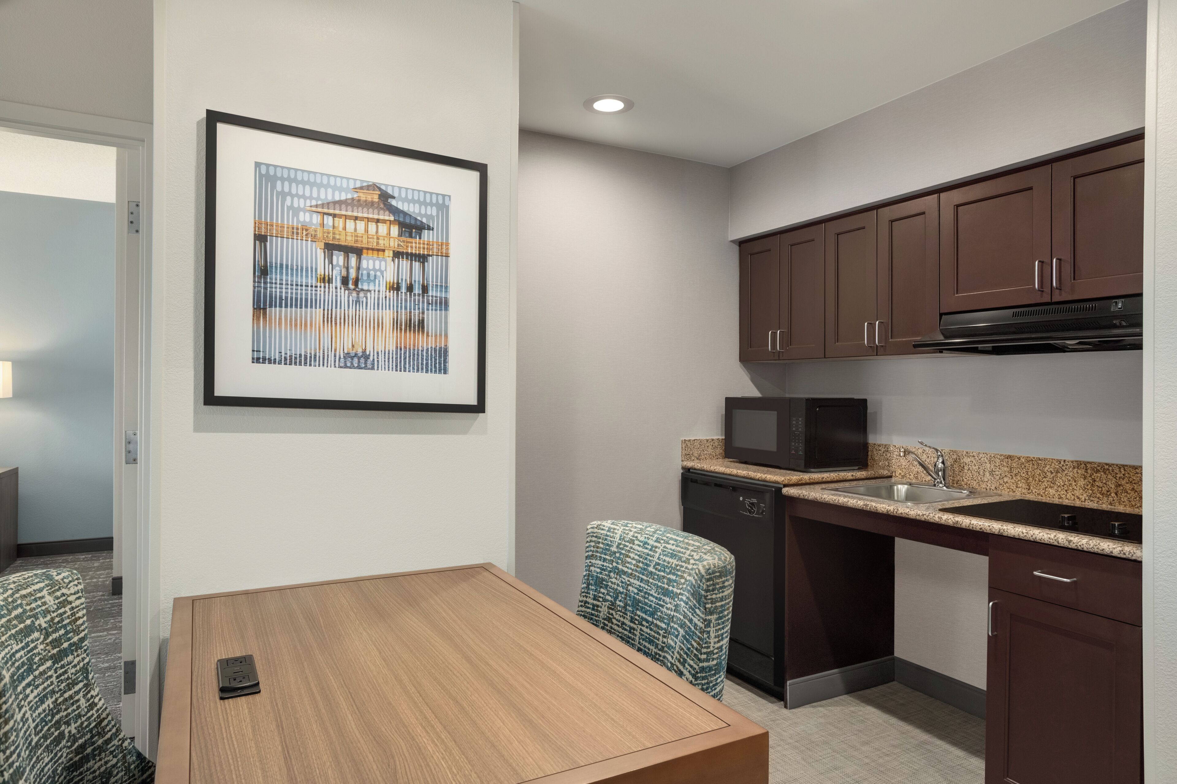 A compact kitchen with a microwave, sink, and dark cabinets. A framed picture hangs on the wall, and a table with a power strip is in the foreground.