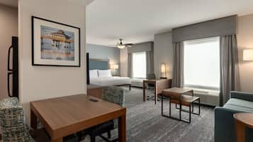A hotel room with a bed, a desk, a chair, a small table, a lamp, and a large window with curtains.