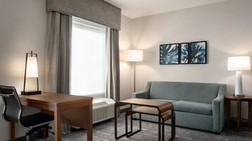 A hotel room with a desk, chair, sofa, and a small table. There are two lamps, a window with curtains, and a wall-mounted picture.
