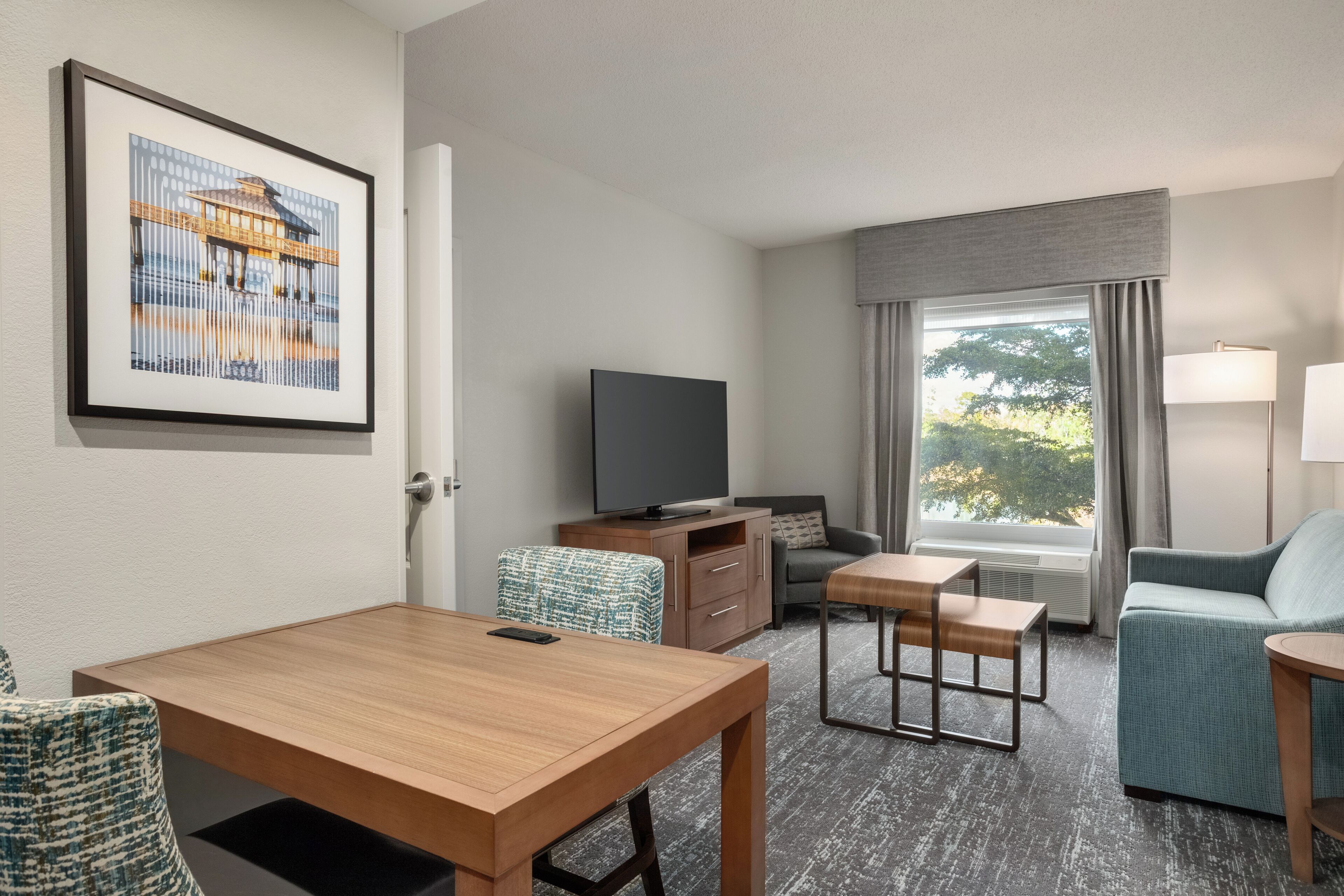 A hotel room with a wooden table, a sofa, a TV, a window with curtains, and a framed picture on the wall.