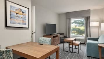 A hotel room with a wooden table, a sofa, a TV, a window with curtains, and a framed picture on the wall.