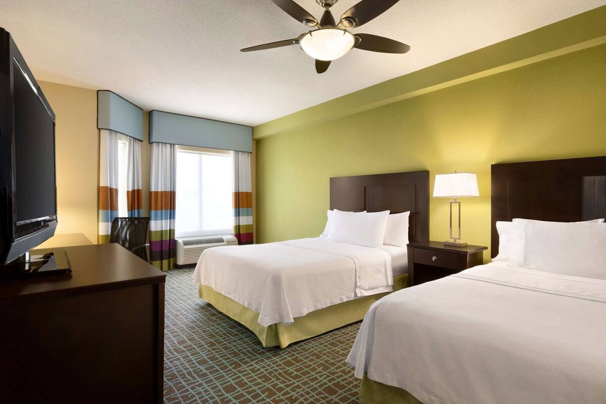 Habitación de hotel con dos camas, un escritorio con televisor, ventilador de techo y ventana con cortinas.