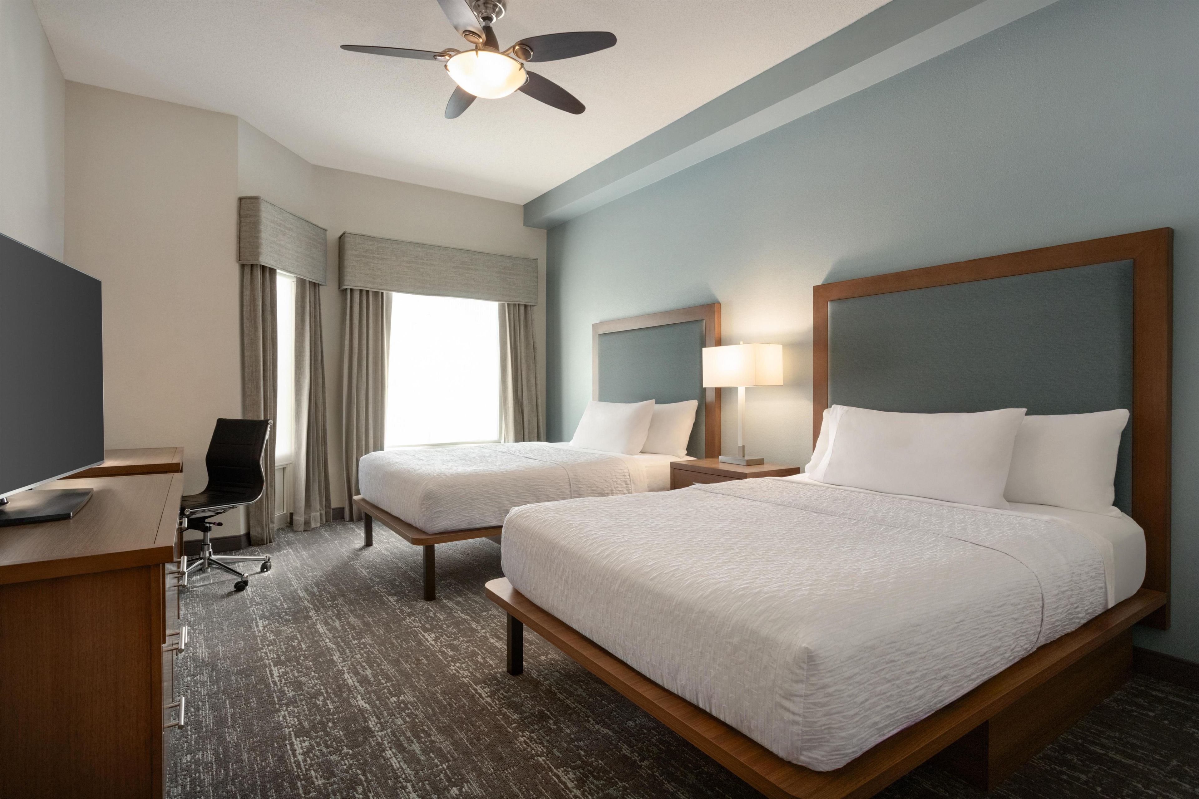 Une chambre d’hôtel avec deux lits, un bureau, une chaise, un ventilateur de plafond et une fenêtre avec des rideaux.