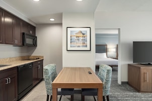 A compact hotel room with a kitchenette, a dining area with two chairs, a bed, a TV, and a framed picture on the wall.