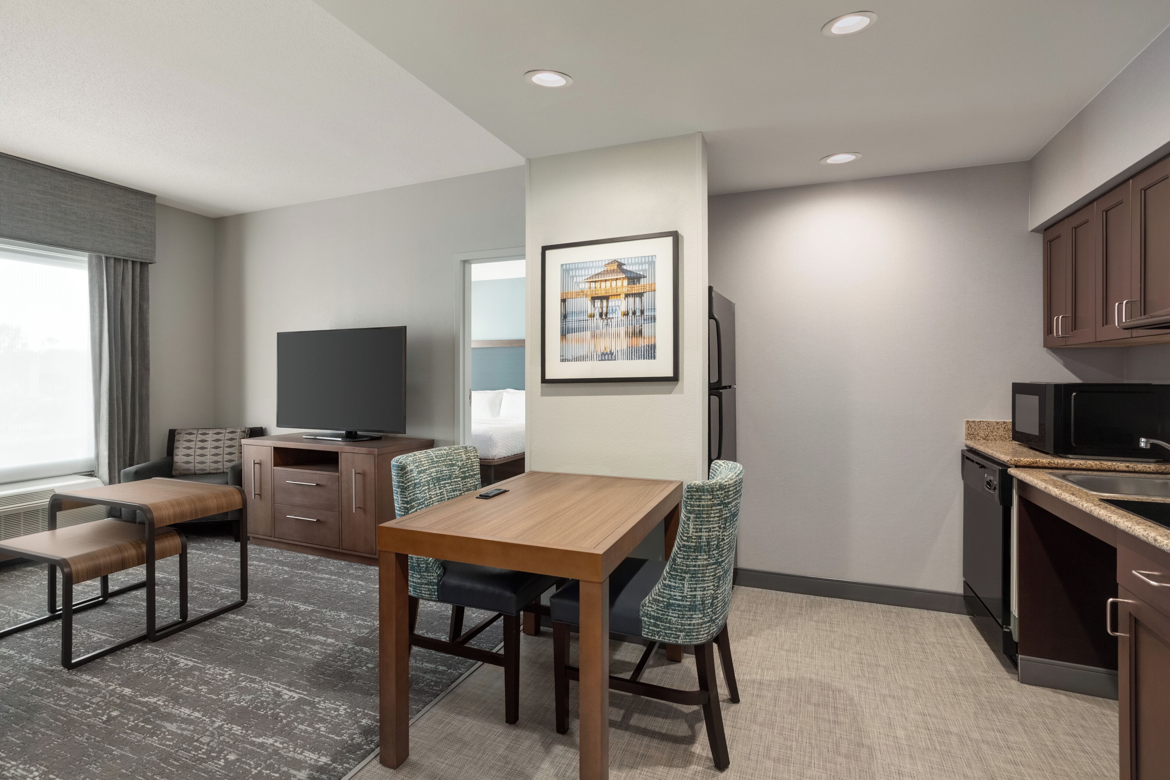 A modern hotel room with a dining area, a flat-screen TV, a kitchen with a microwave, and a framed picture on the wall.