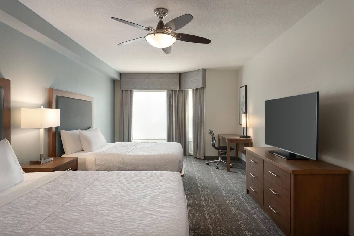 Habitación de hotel con dos camas, ventilador de techo, televisor, escritorio y una silla.