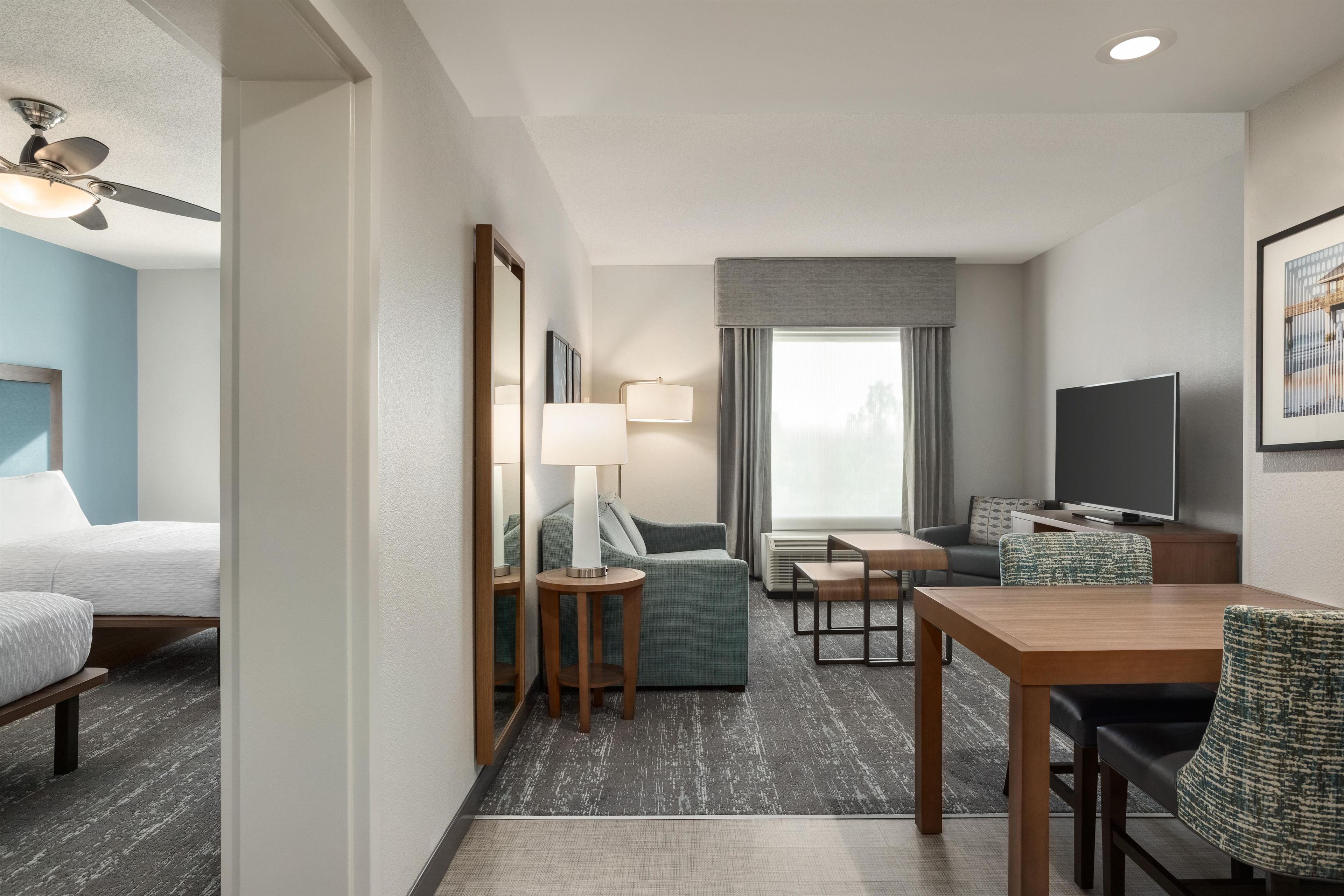 A hotel room with a bed, a desk with a chair, a television, a window with curtains, and a ceiling fan.