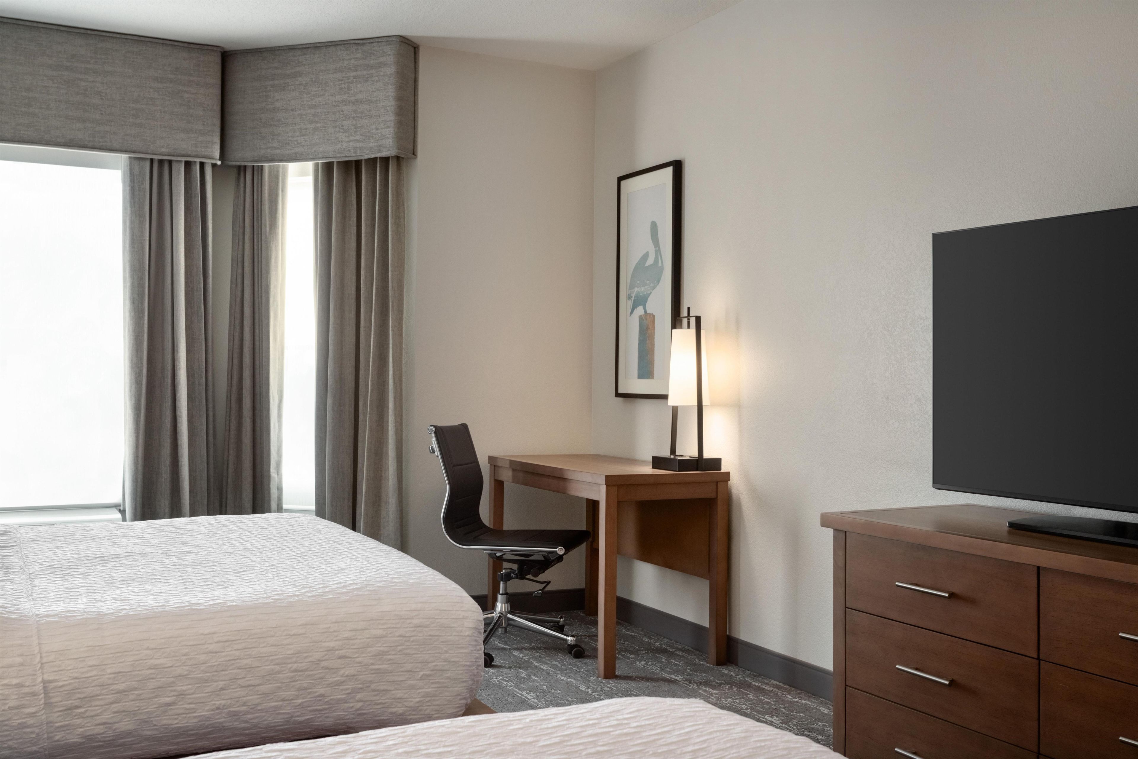 Quarto de hotel com duas camas, uma escrivaninha, uma cadeira, uma televisão e um quadro na parede.