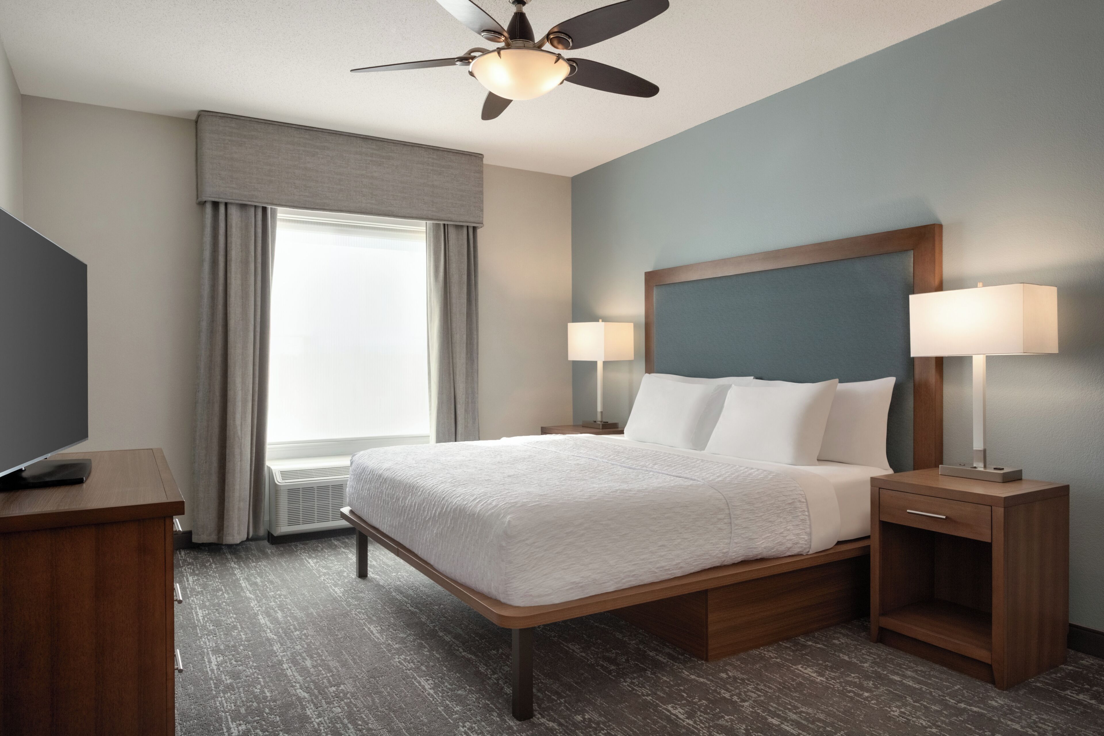 A hotel room with a bed, two bedside tables with lamps, a ceiling fan, a window with curtains, and a TV on a stand.