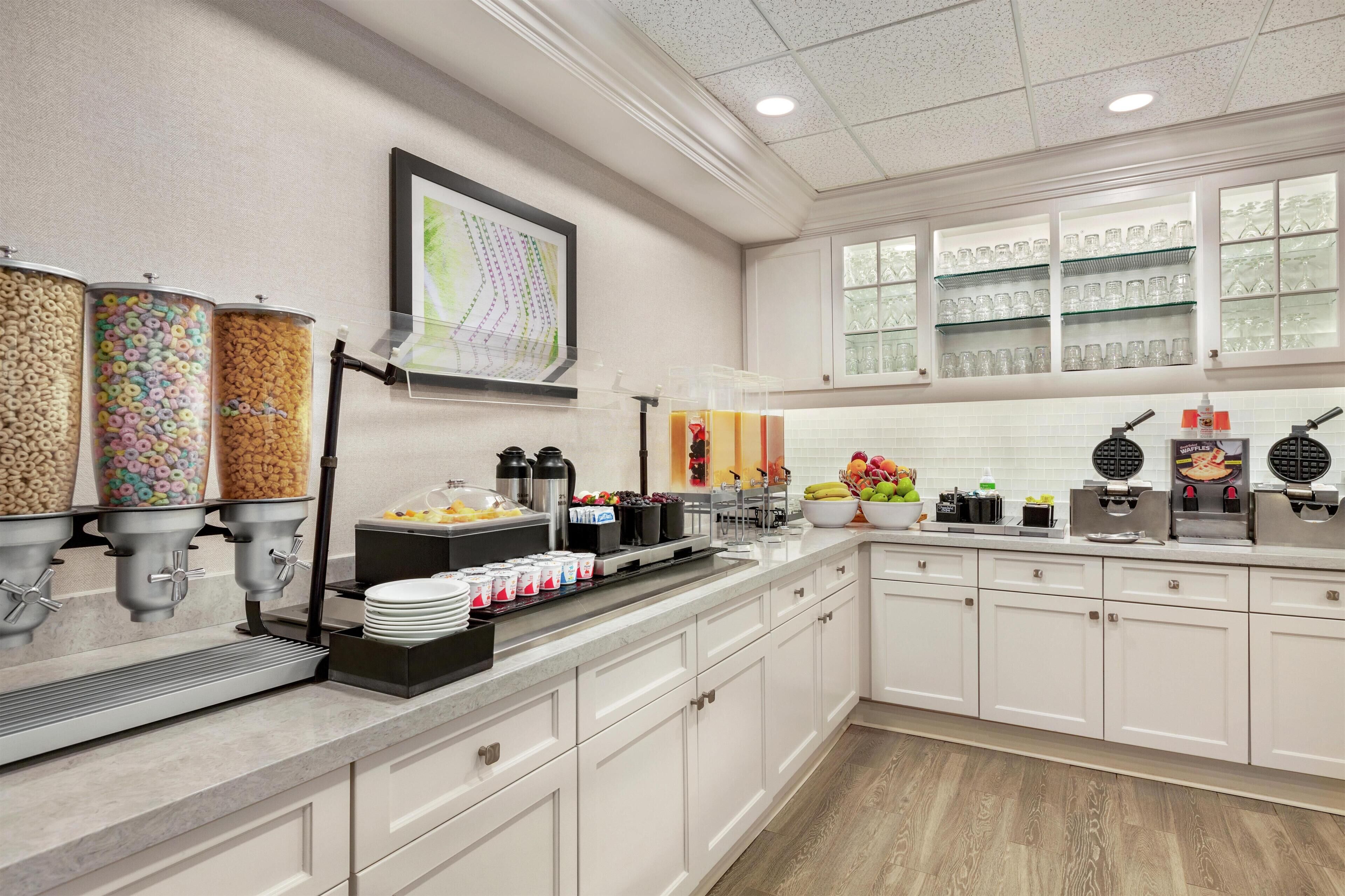 A modern kitchen with a breakfast bar, cereal dispensers, a fruit bowl, and a waffle maker.