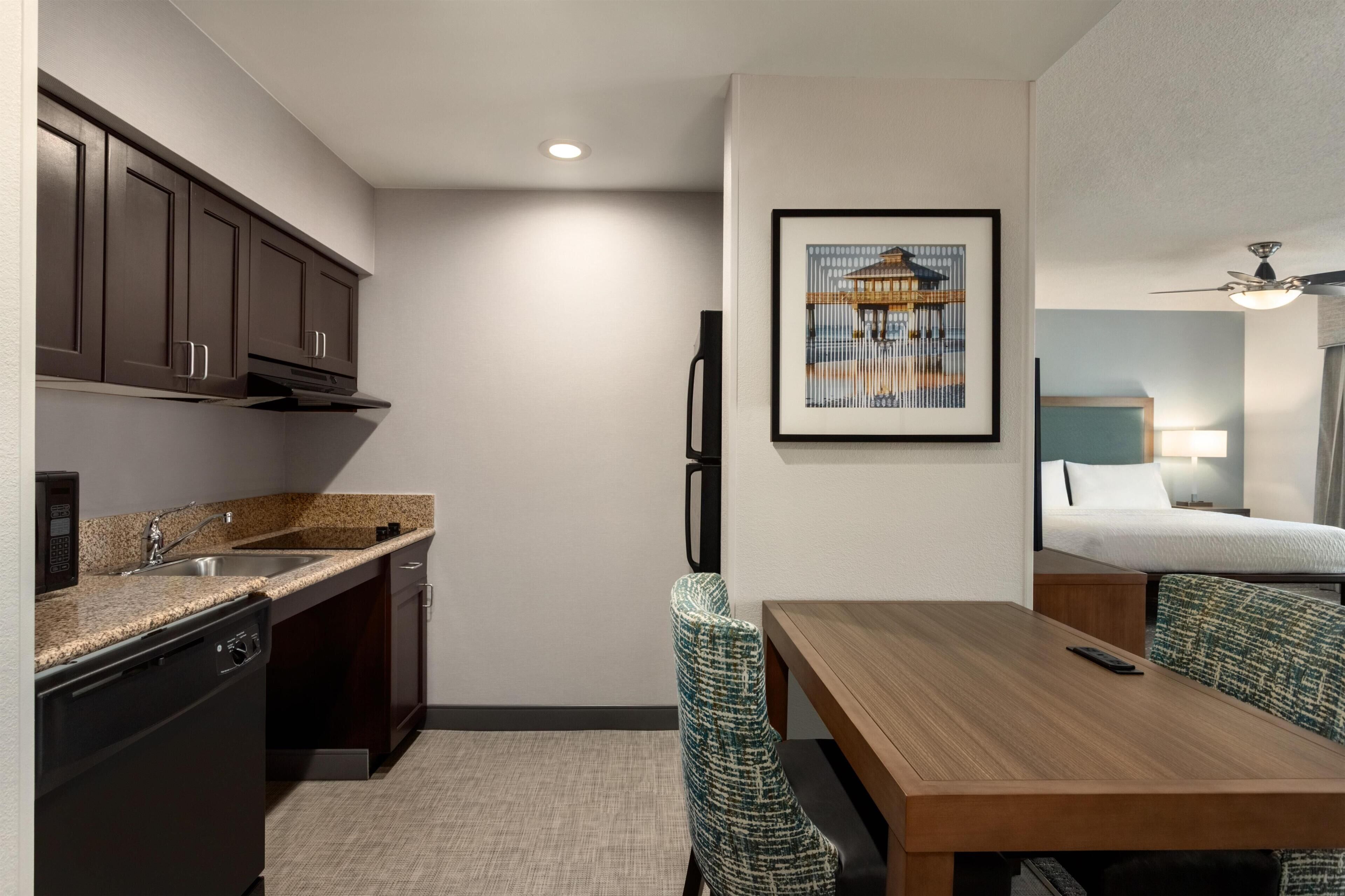 A compact kitchen with a dishwasher, a sink, and a microwave. There is a dining area with a wooden table and two chairs. A bed with a headboard and a nightstand with a lamp is visible in the background.