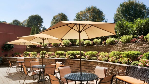 Een buitenterrein met ronde tafels en stoelen, beschaduwd door grote parasols, omringd door een aangelegde tuin met diverse planten en een stenen muur.