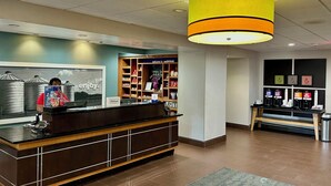 Zona de recepción de un hotel con un mostrador de madera, una lámpara colgante amarilla grande, un cuadro en la pared y una estación de café con varios dispensadores de bebidas.