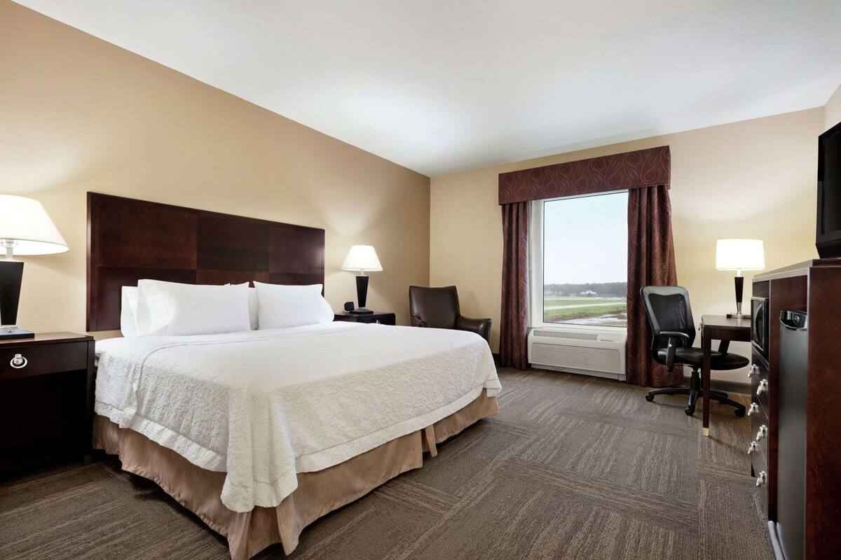 A hotel room with a large bed, two bedside tables, a desk with a chair, a television, and a window with curtains.