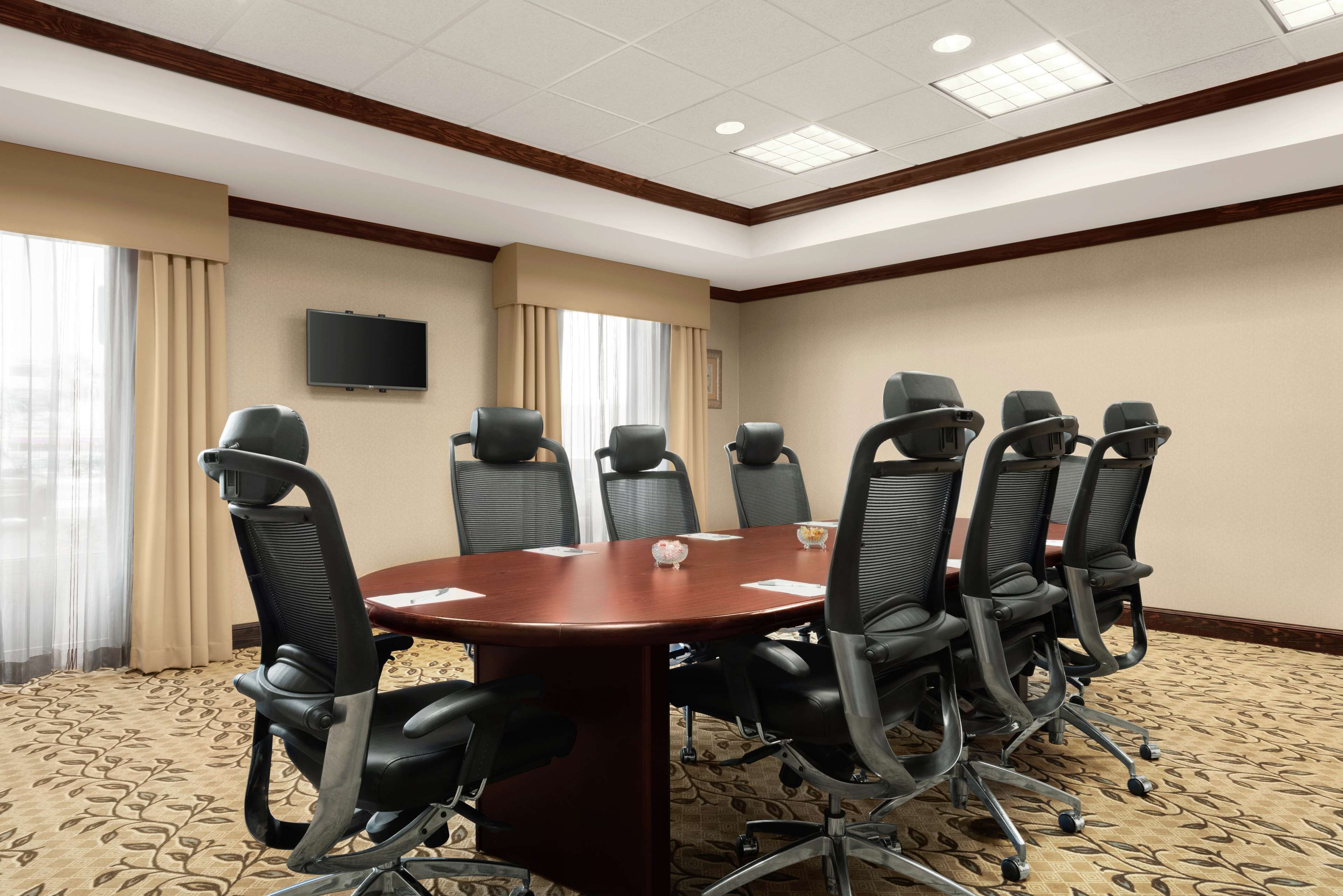 A conference room with a large table, eight chairs, a television on the wall, and large windows with curtains.