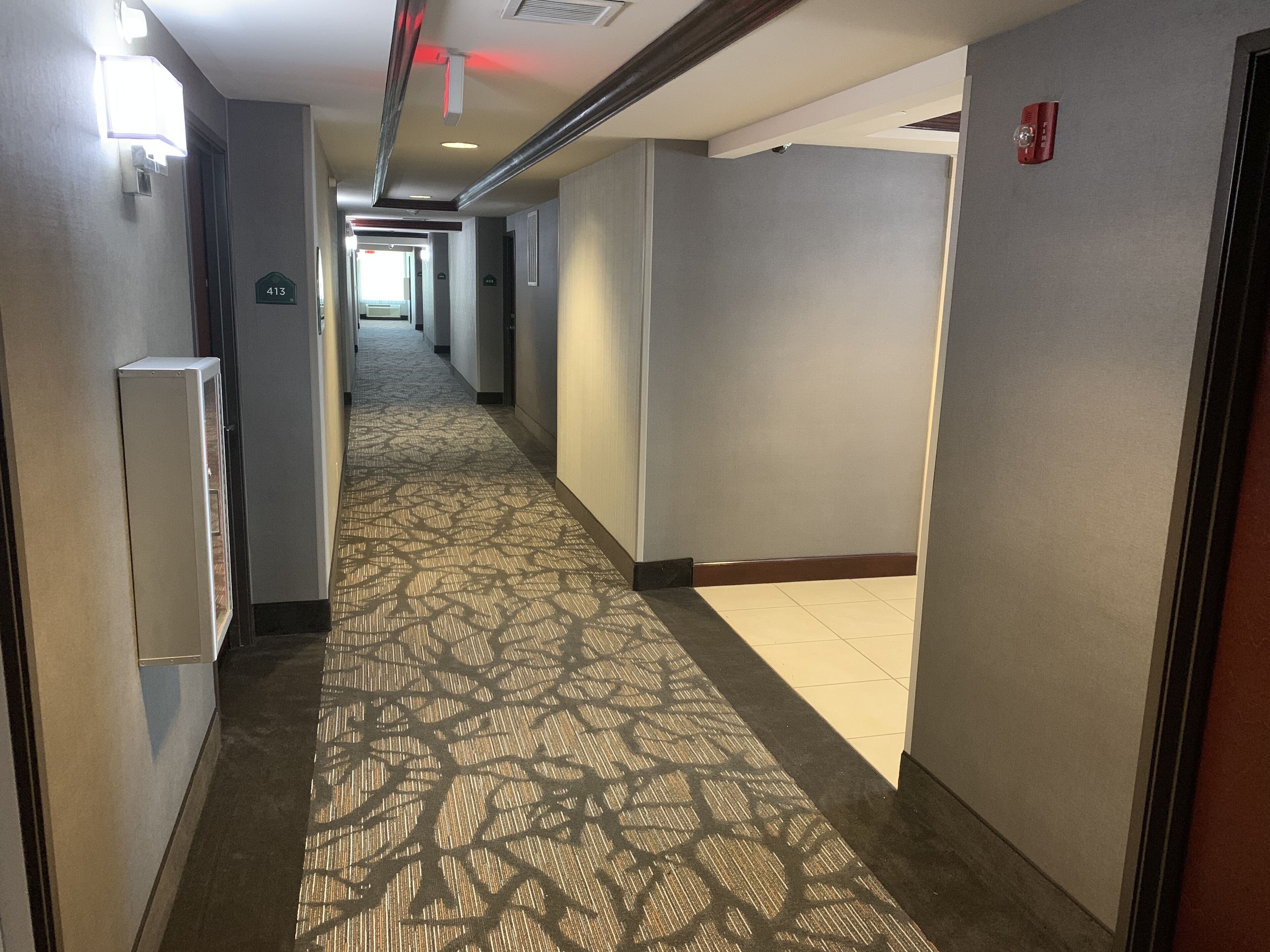 Pasillo de hotel con alfombra estampada, un detector de incendios en la pared y una puerta con un letrero de número.