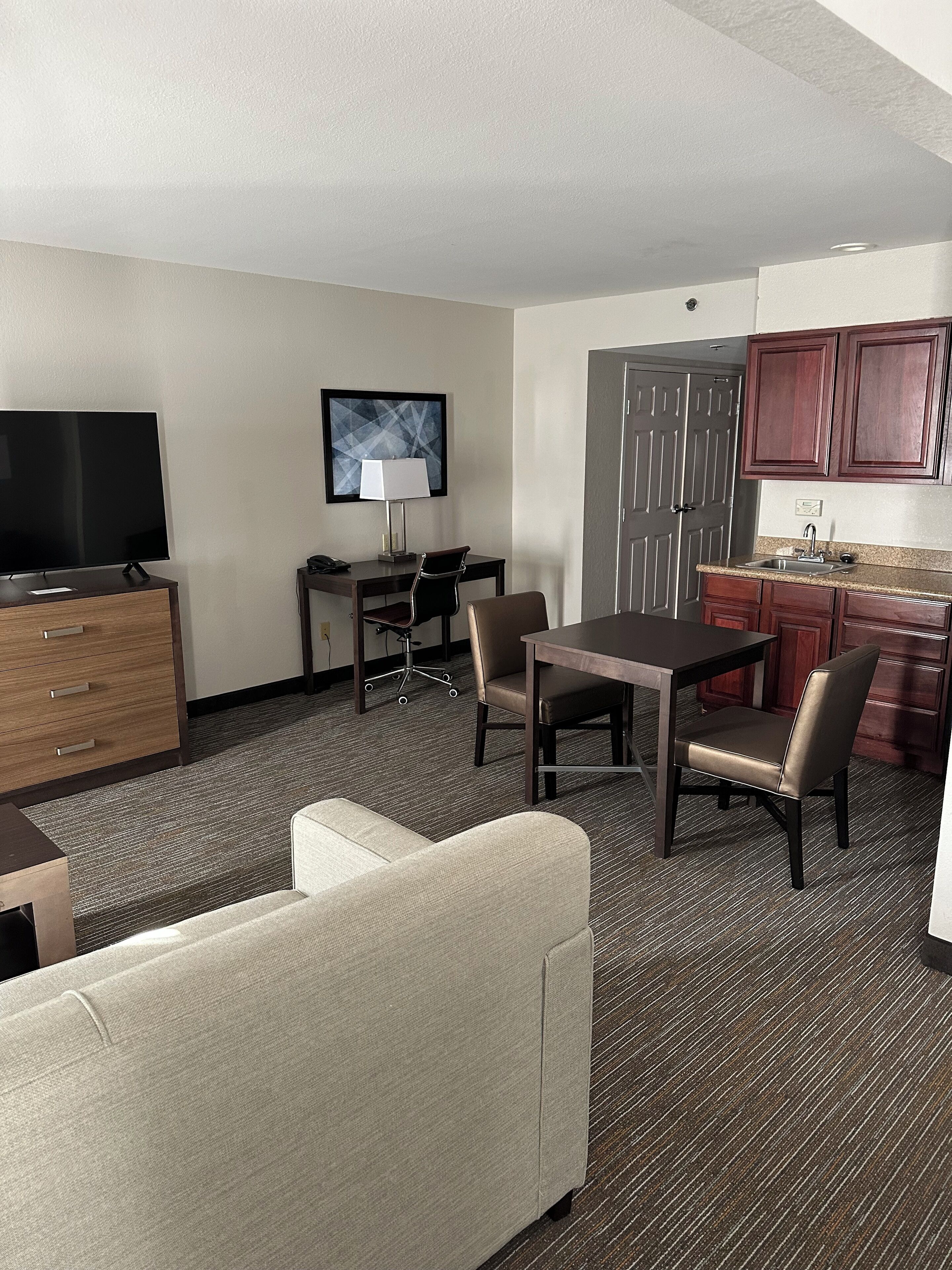 Habitación de hotel con televisor de pantalla plana, escritorio con lámpara, mesa de comedor con sillas, sofá y kitchenette con armarios.