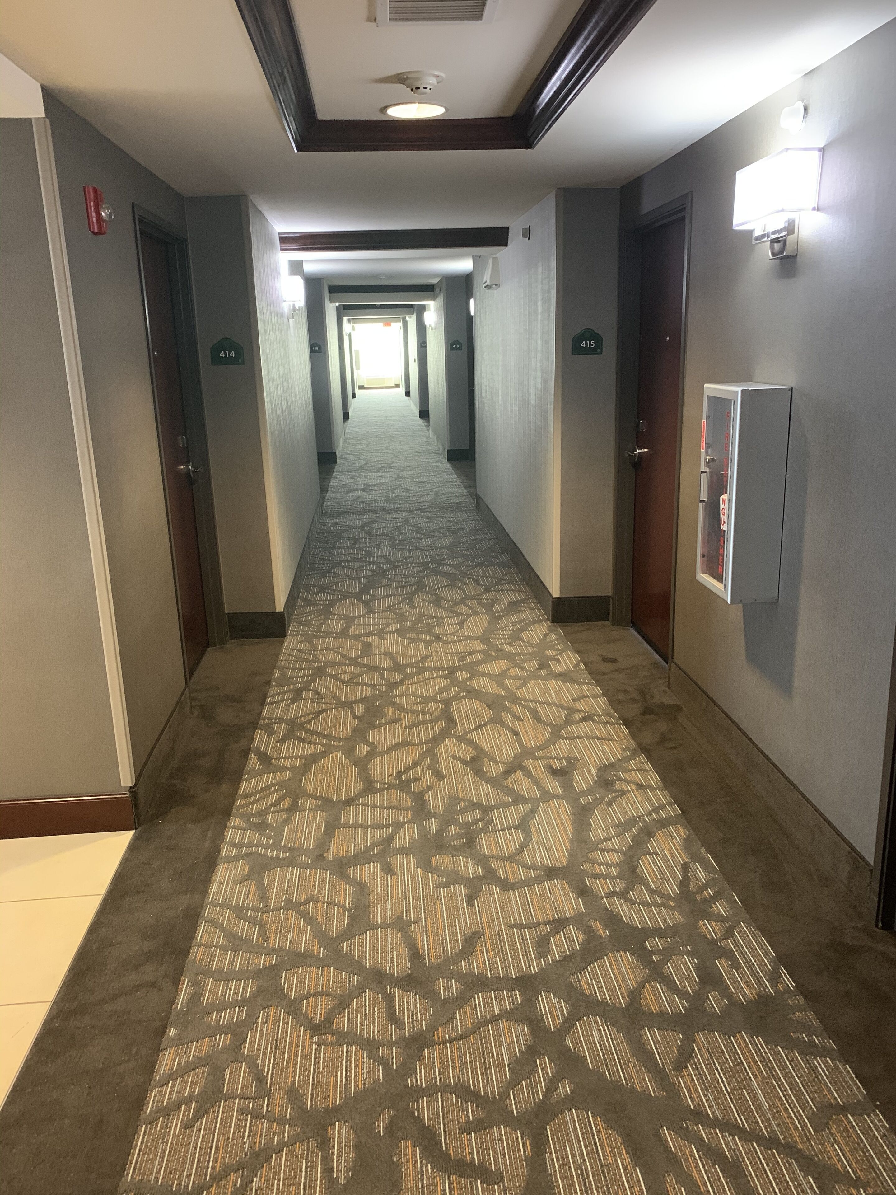 A hotel hallway with carpeted floors, doors marked with room numbers, and a light fixture on the wall.