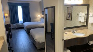 A hotel room with two beds, a large window with blue curtains, a sitting area with a chair and table, and a bathroom with a sink and mirror.