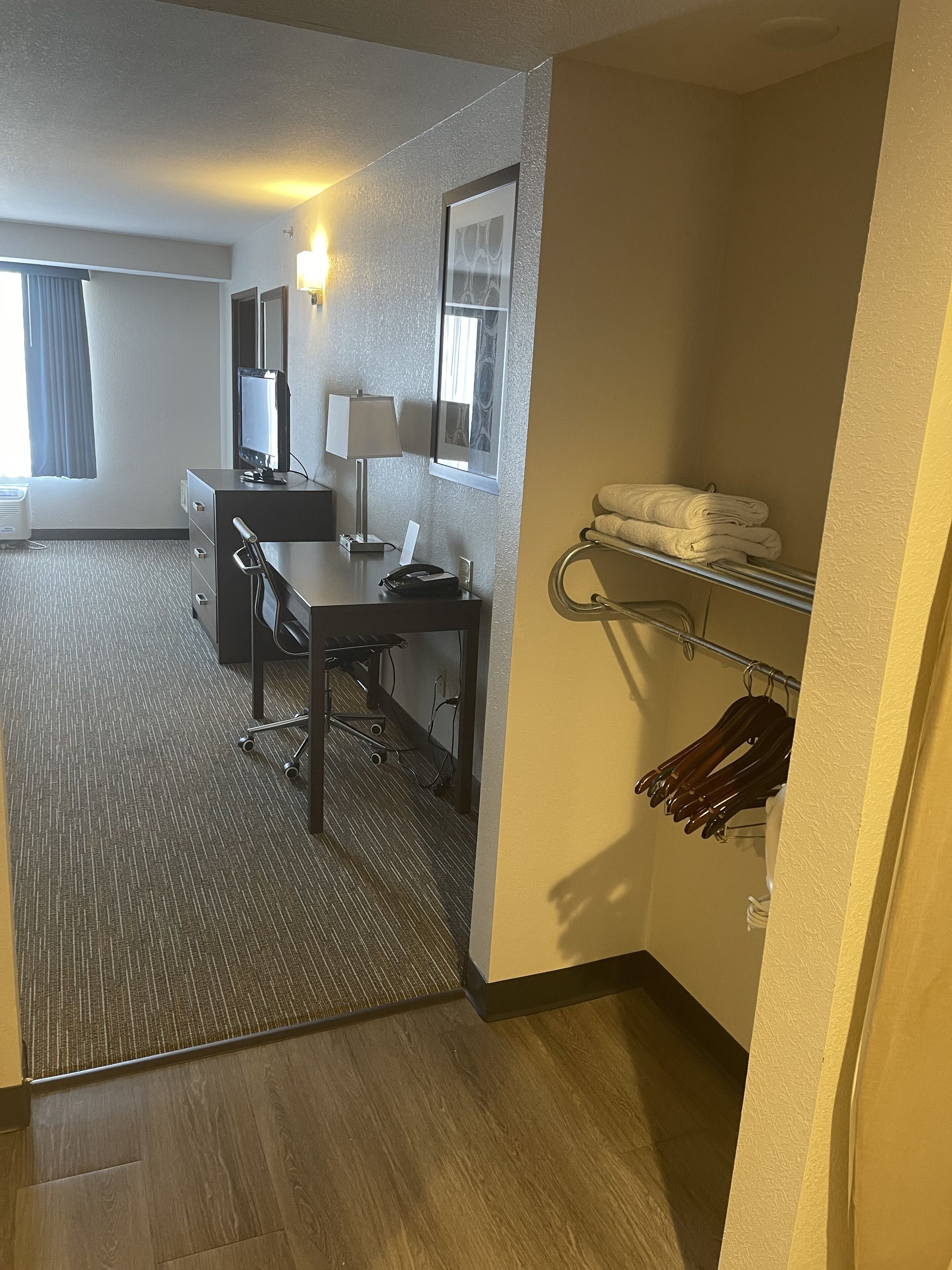 Une chambre d’hôtel équipée d’un bureau, d’une chaise et d’une petite kitchenette. On y trouve une fenêtre avec des rideaux, un téléviseur fixé au mur et un porte-serviettes avec des serviettes.