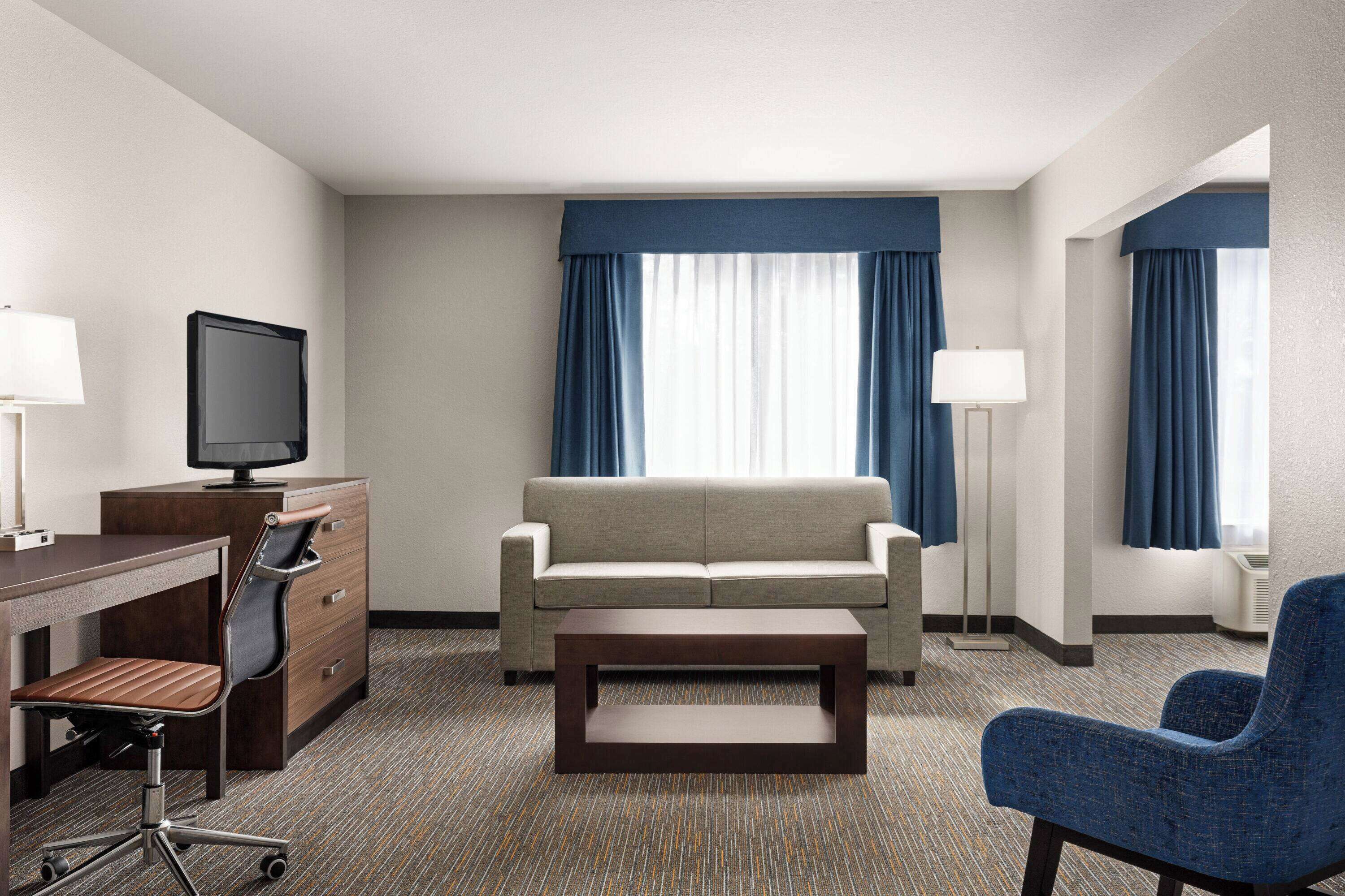 A hotel room with a sofa, a desk with a chair, a television, and a lamp.