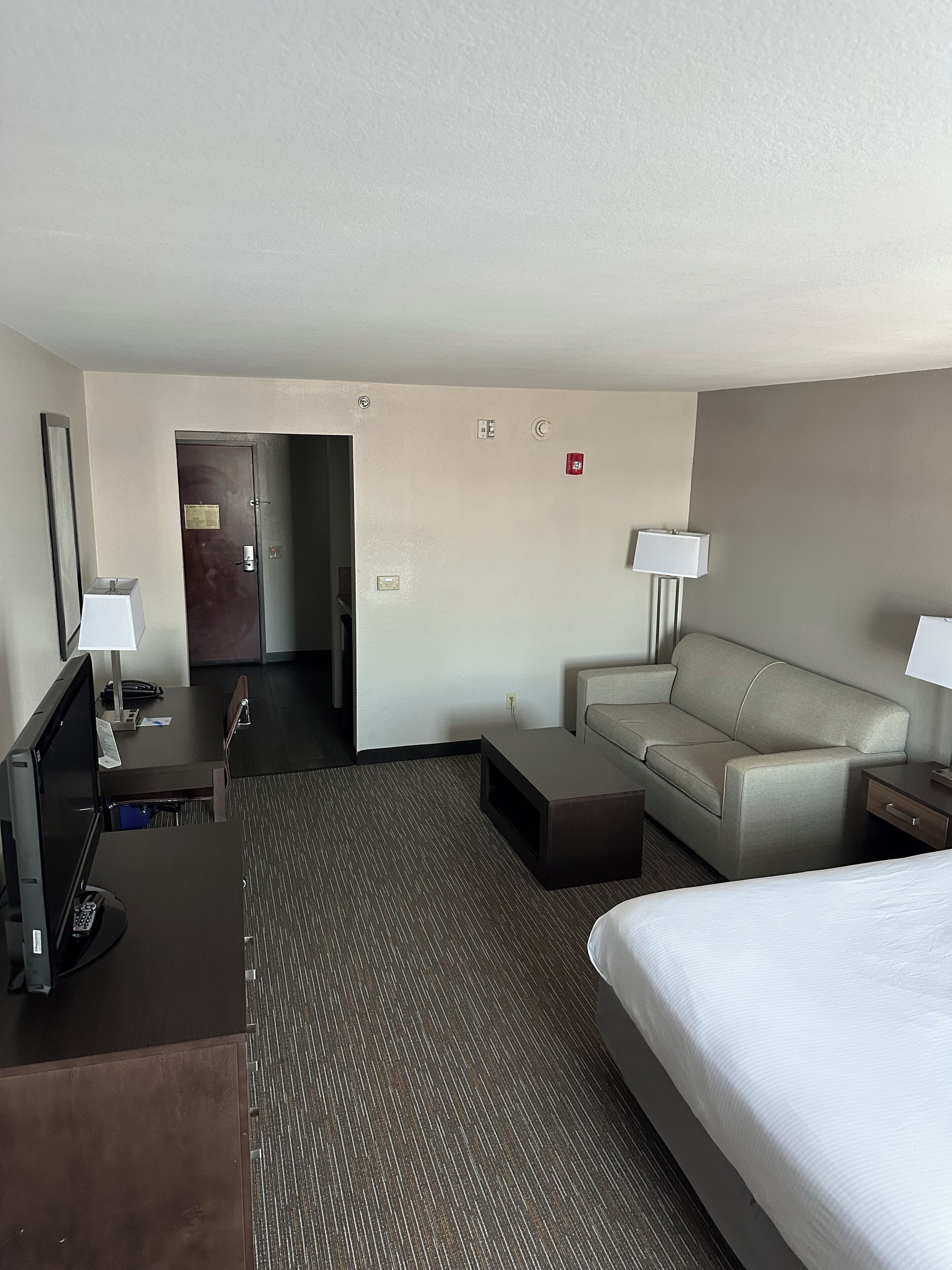 Une chambre d’hôtel avec un lit, un bureau équipé d’un ordinateur, un canapé et une petite table. Il y a une porte qui donne sur une autre pièce et un détecteur d’incendie au mur.