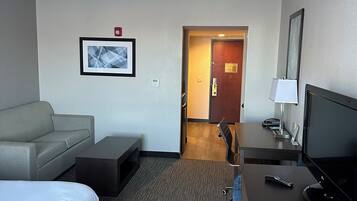 Een hotelkamer met een bed, een bank, een bureau met een televisie, een stoel en een deur die naar een andere kamer leidt.