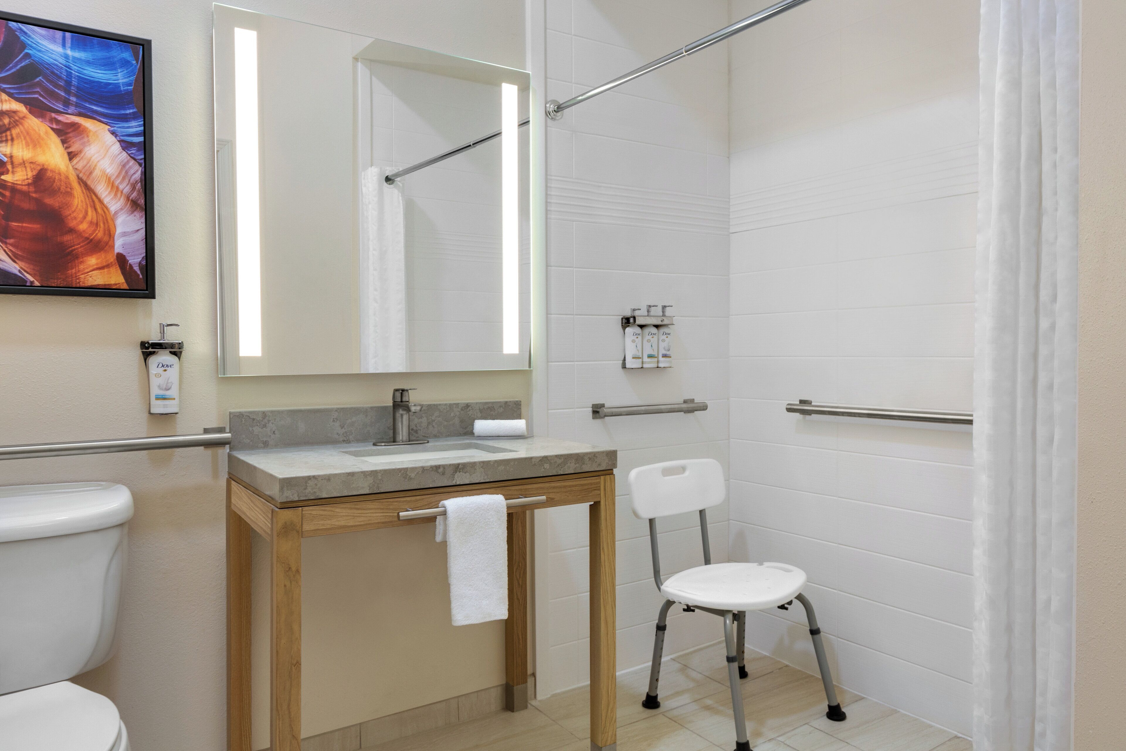 A bathroom with a toilet, sink, mirror, shower, and a chair.