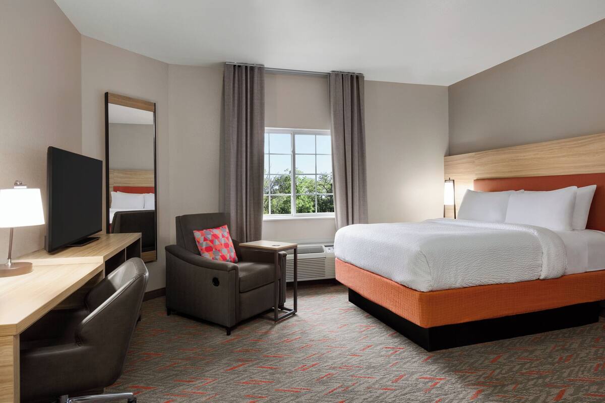Una habitación de hotel con cama, escritorio con televisor, silla, ventana con cortinas y suelo alfombrado.