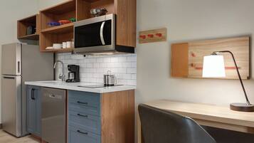 A compact kitchen with a microwave, coffee maker, and a wooden shelf with dishes.