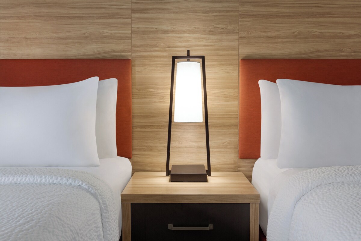 Una habitación de hotel con dos camas, una mesita de noche con lámpara y un cabecero de madera.