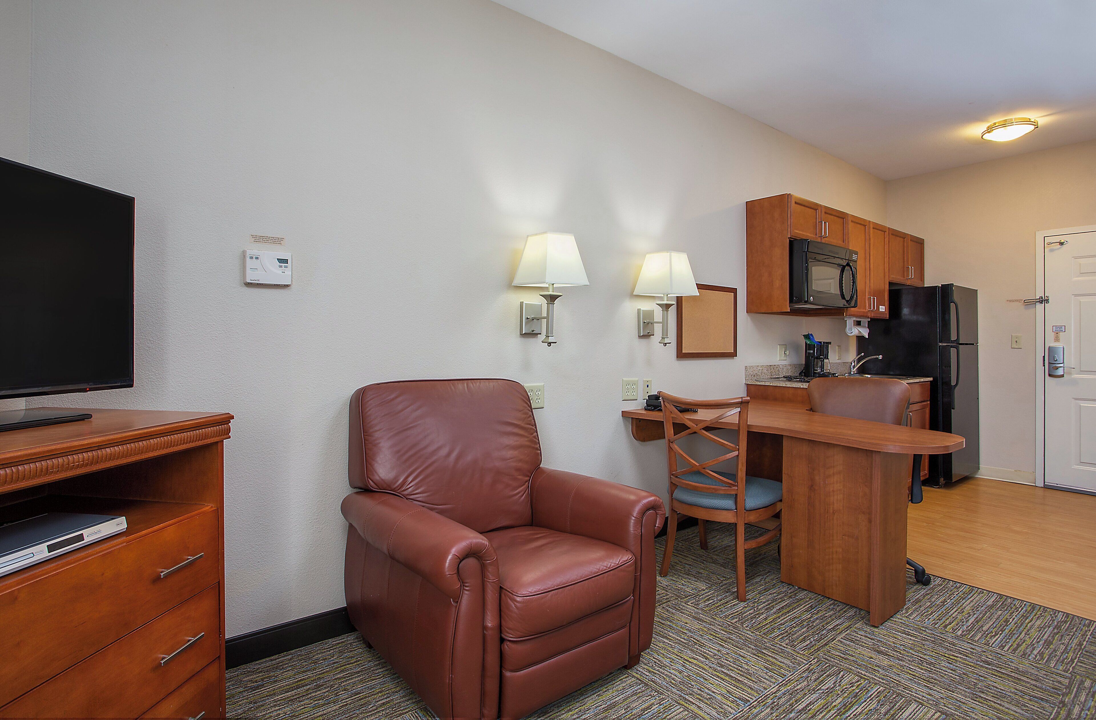A compact hotel room with a television, a leather chair, a small desk with a chair, a microwave, and a refrigerator.