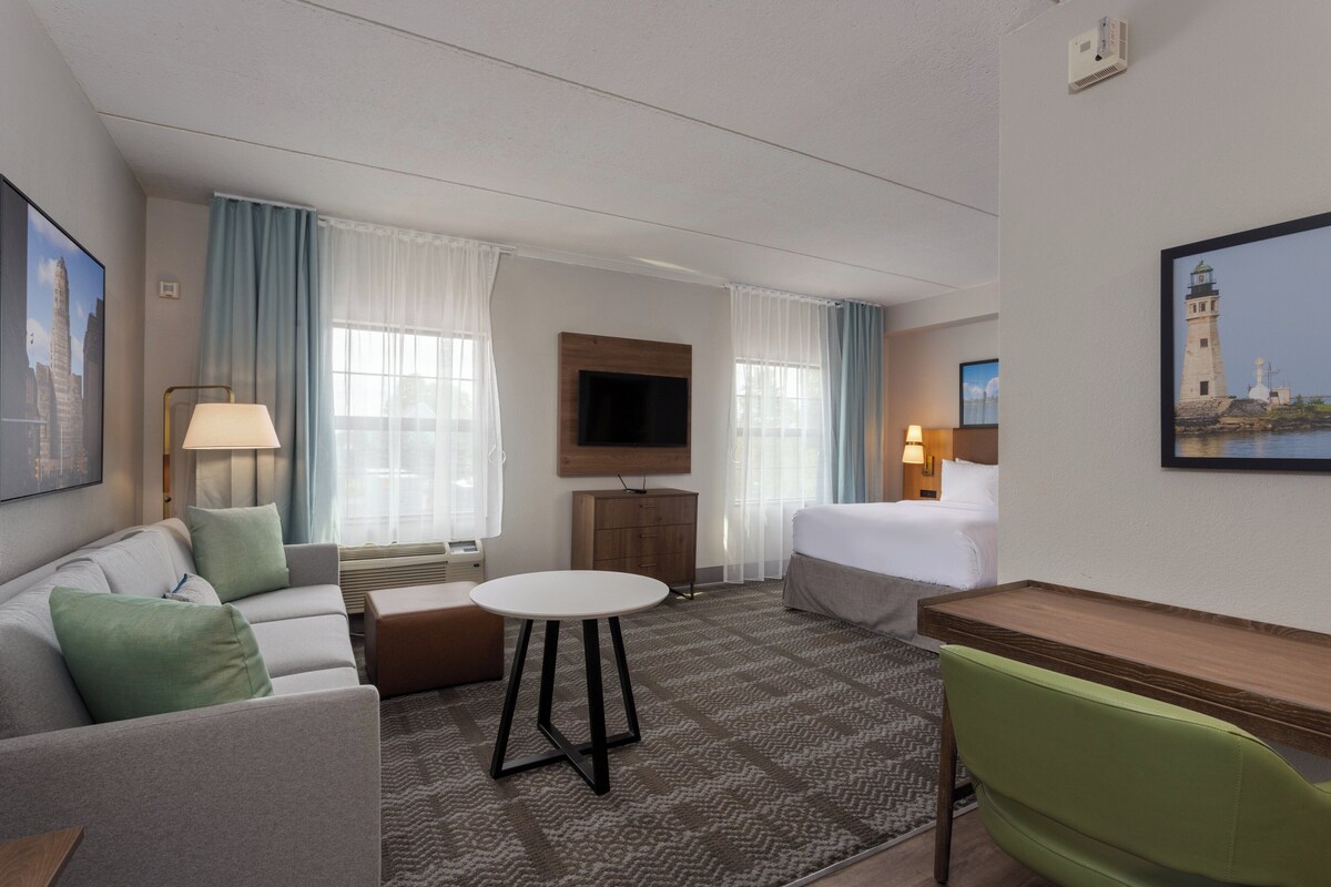 Een hotelkamer met een bed, een bureau, een bank, een televisie en een schilderij van een vuurtoren.