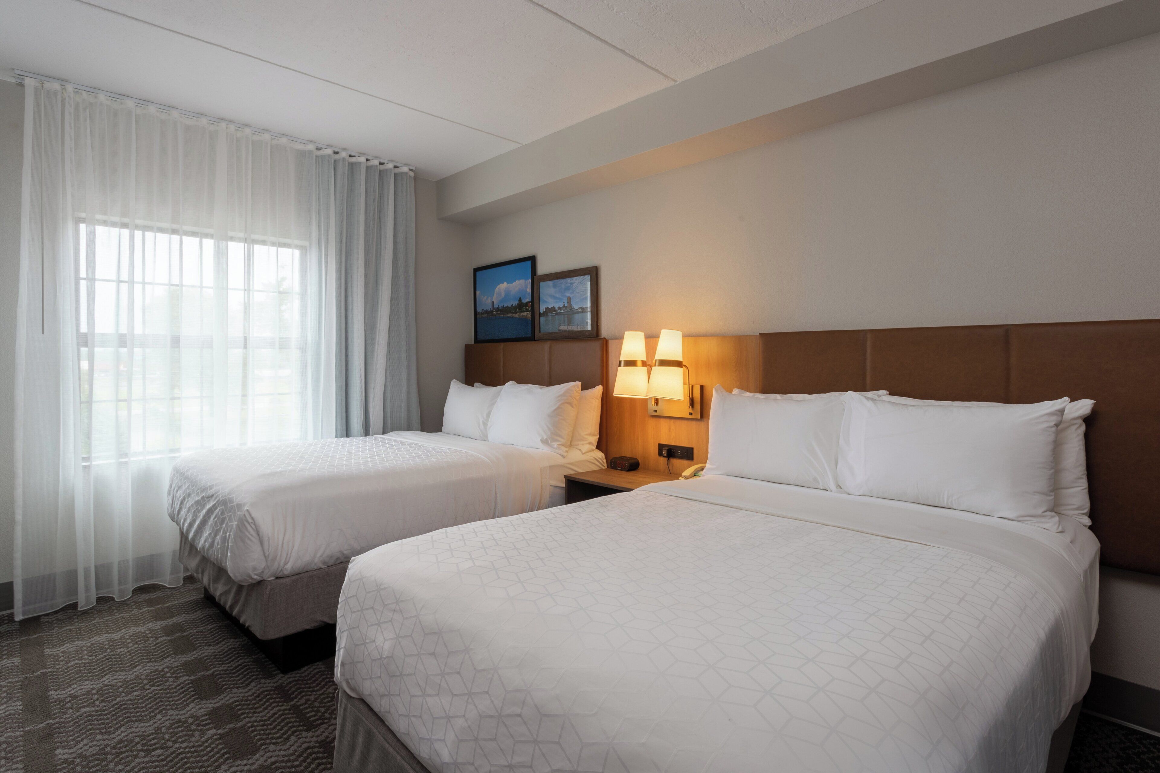 A hotel room with two beds, a large window with sheer curtains, a nightstand with a lamp, and two framed pictures on the wall.