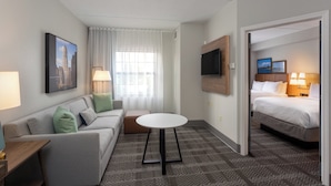 A hotel room with a bed, a sofa, a small table, a TV, and a window with curtains. - Staybridge Suites West Seneca by IHG (Buffalo)