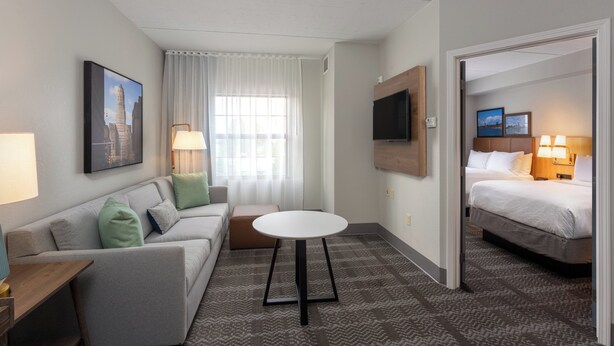 A hotel room with a bed, a sofa, a small table, a TV, and a window with curtains. - Staybridge Suites West Seneca by IHG (Buffalo)