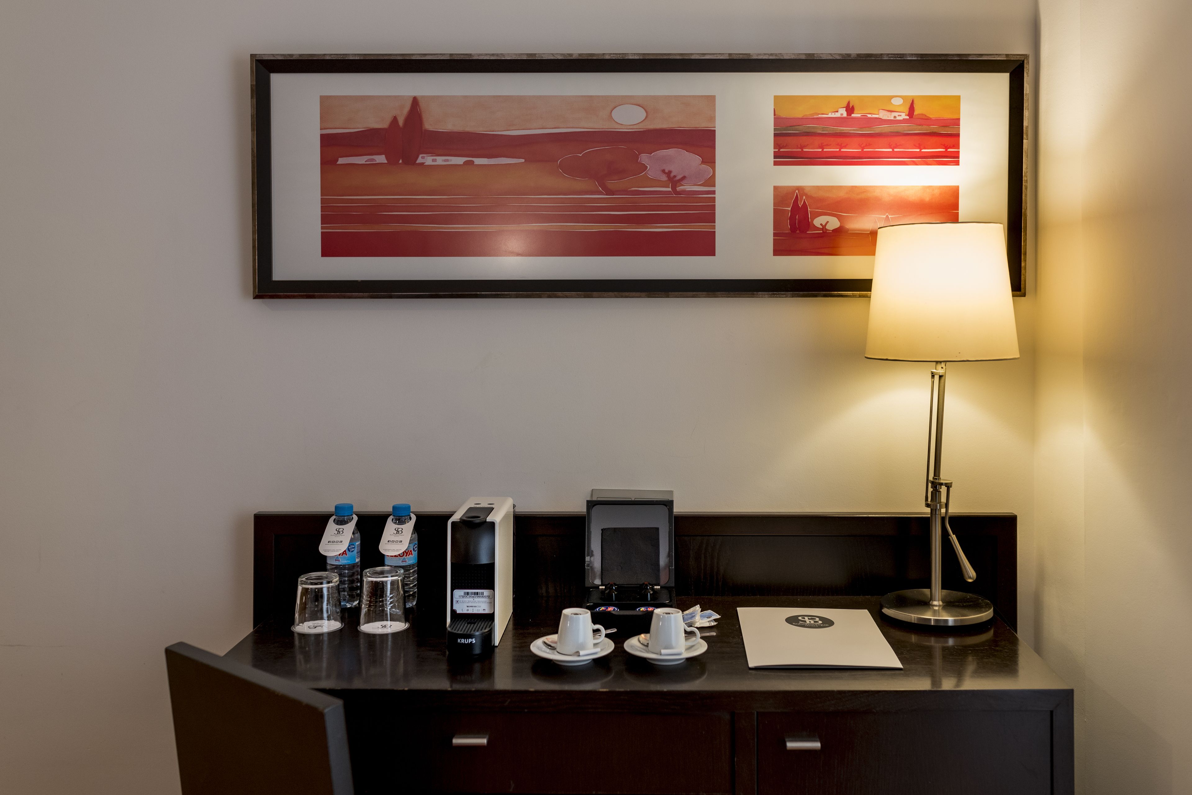 Un comptoir d’accueil d’hôtel avec une œuvre d’art encadrée au-dessus, une lampe de bureau et une machine à café.