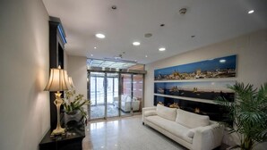 A modern lobby with a white sofa, a black console table with a lamp, and a large painting of a coastal cityscape. - Hotel ITC Salamanca by Soho Boutique (Salamanca)