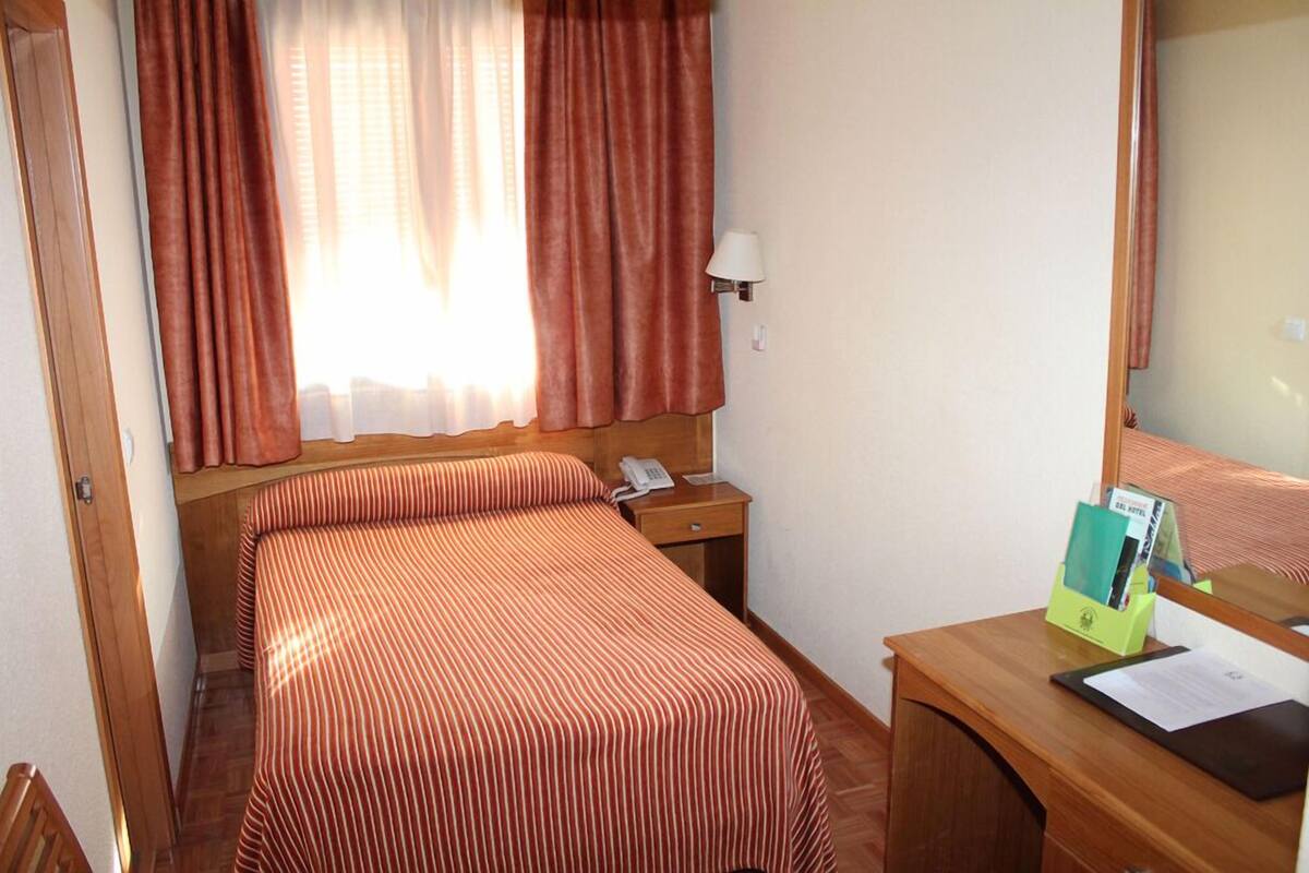 Ett hotellrum med en säng, ett sängbord med en lampa, en spegel och ett skrivbord med en stol.