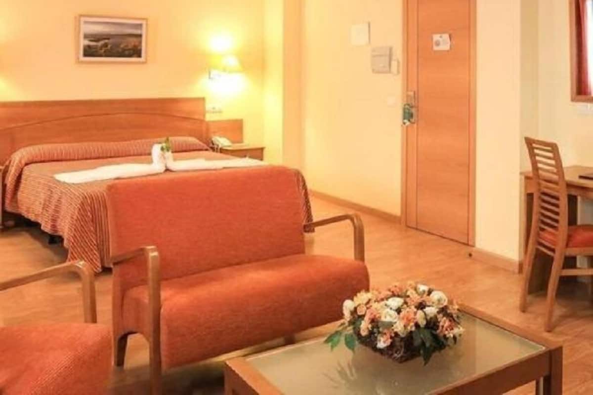 Ett hotellrum med två sängar, en sänggavel i trä, ett skrivbord med en stol, ett litet bord med en blomsteruppsättning och en vägglampa.