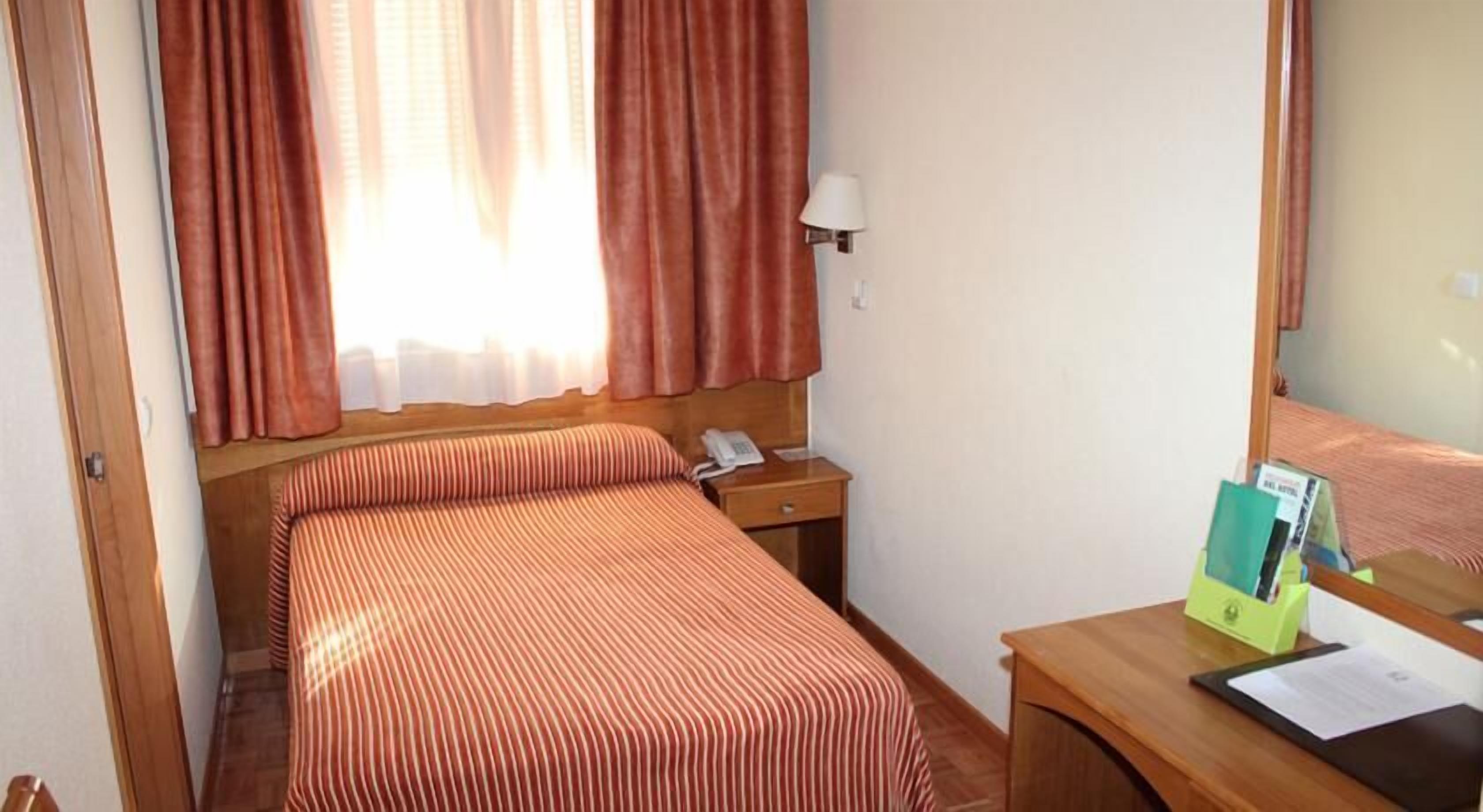 A hotel room with a bed, a bedside table with a lamp, a telephone, and a desk with a mirror and some papers.