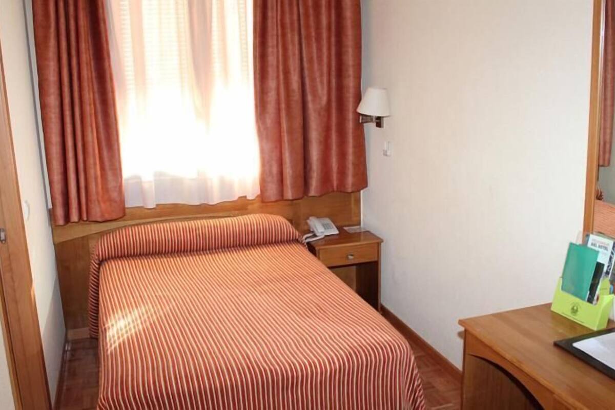 Ett hotellrum med en säng, ett nattduksbord med en lampa, en telefon och ett skrivbord med en spegel och några papper.