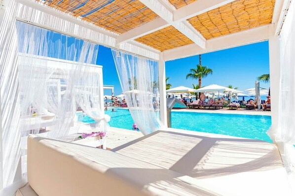 A poolside lounge with white canopies, a pool, and palm trees in the background.