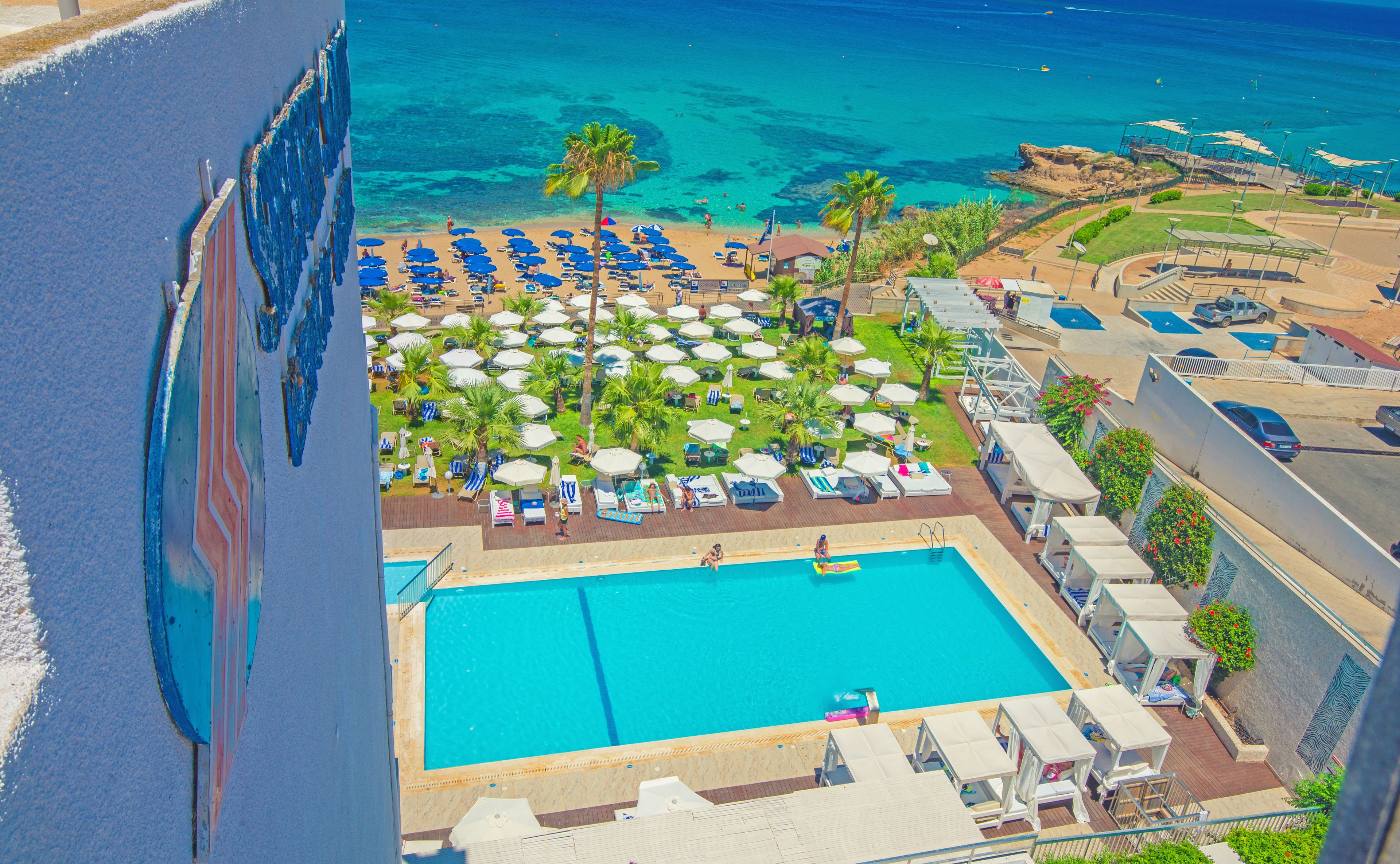 Vista aérea de un complejo turístico con una piscina grande, varios sillones de playa y sombrillas.