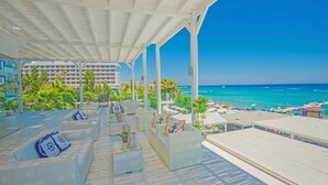 A covered outdoor seating area with wicker furniture and cushions, overlooking a beach with umbrellas and people. - Silver Sands Beach Hotel (Paralimni)