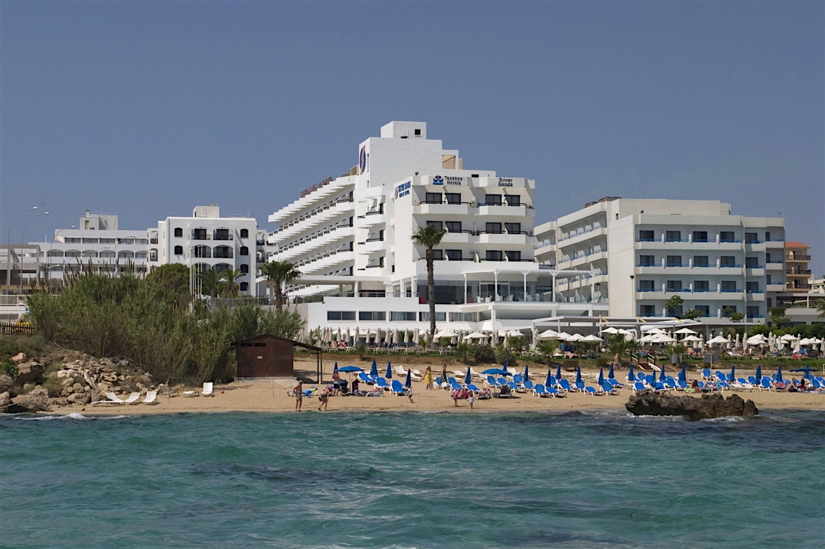 A hotel with multiple floors, a beach with blue umbrellas, and a rocky shoreline.