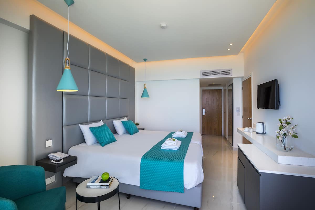 A modern hotel room with a large bed, a desk, a chair, a TV, and a small table with a book and a plant.