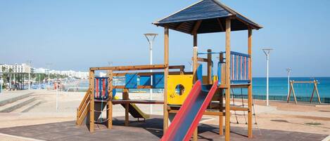 A playground with a slide and climbing structures, located near a beach with a clear view of the sea.