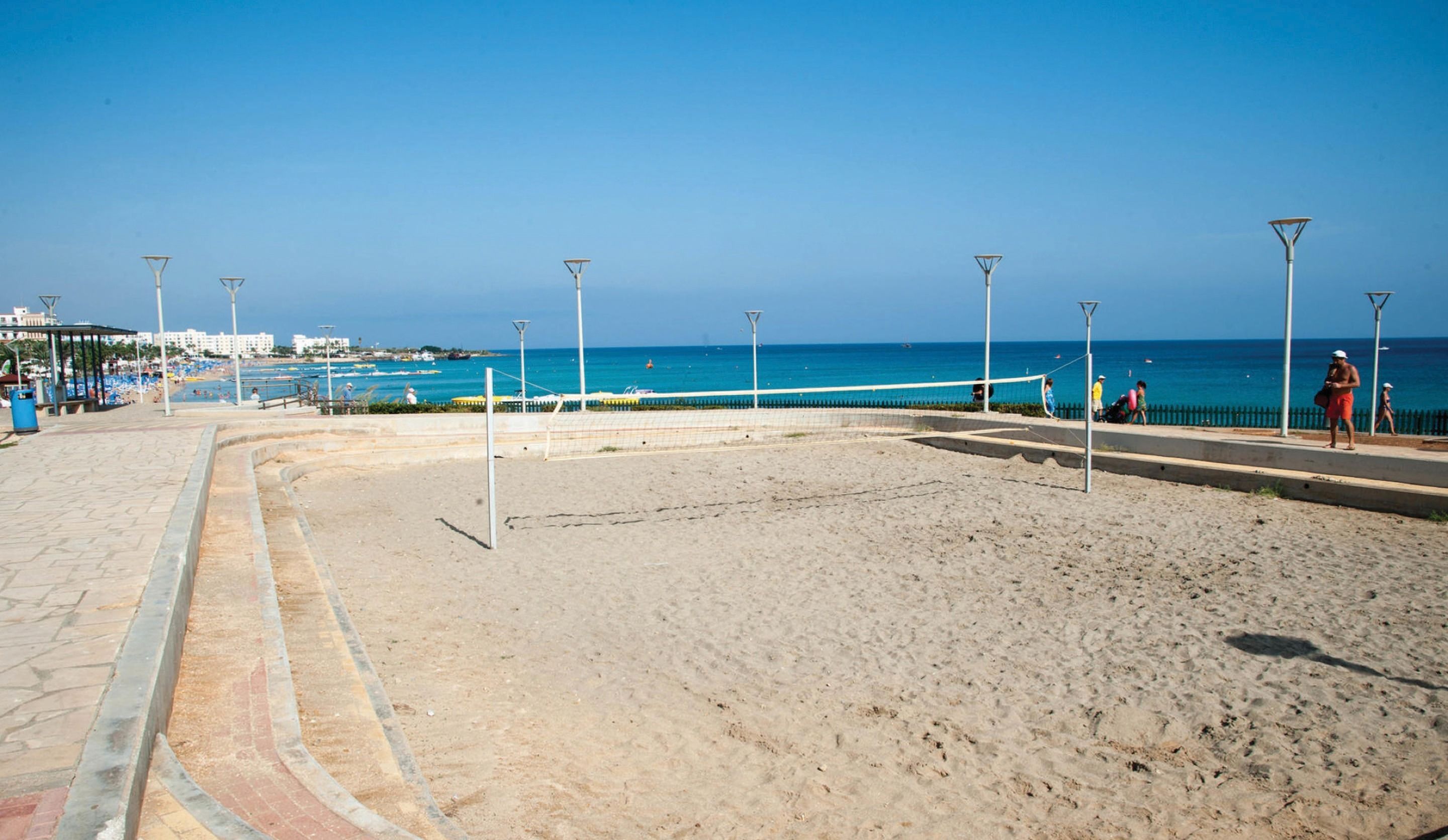 A beach volleyball court with a net, a paved walkway, and a fence.
