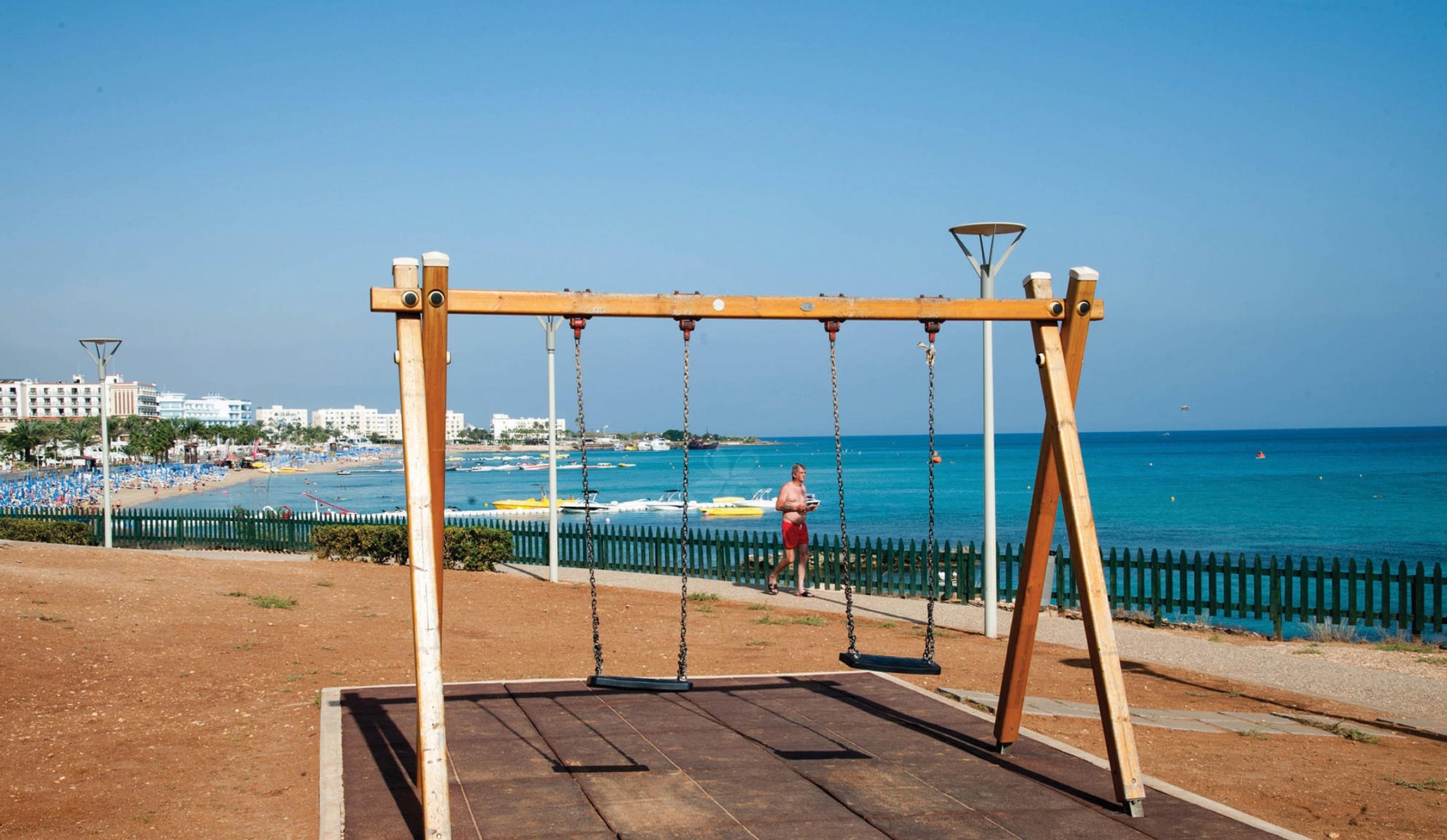 Un columpio de madera con estructura metálica, cerca de una playa con valla y vista al mar.
